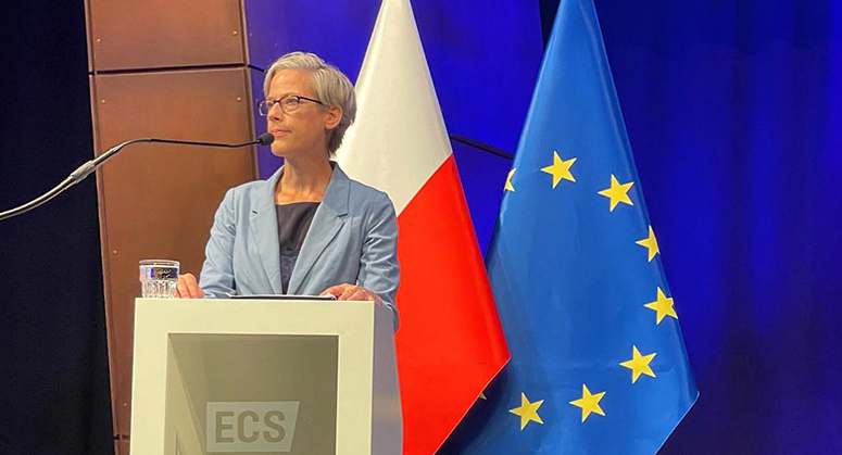 Meg Satterthwaite speaking at a podium with EU flag behind her