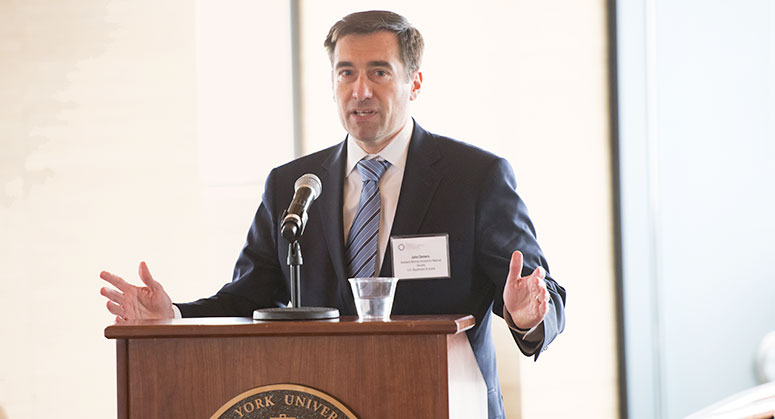 John Demers standing at a podium, addressing the crowd