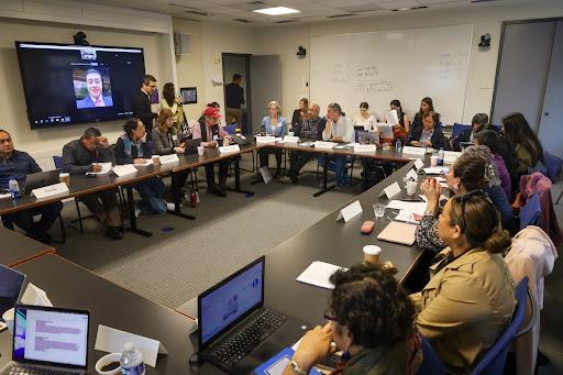 15 people sitting on tables arranged in a hallow square configuration. People are taking notes on their computer and on paper. There is a large TV in the background with an ongoing Zoom call. 